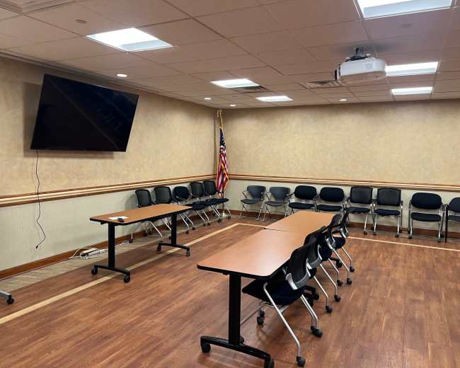 The image shows a meeting room with a long table surrounded by black chairs, a large screen mounted on the wall, and an American flag in the corner.
