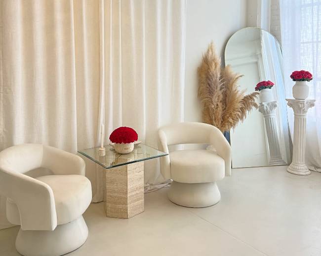 The image shows a bright, minimalistic space with two white chairs, a glass table, and decorative elements like a chandelier, a vase with red flowers, and tall pampas grass.