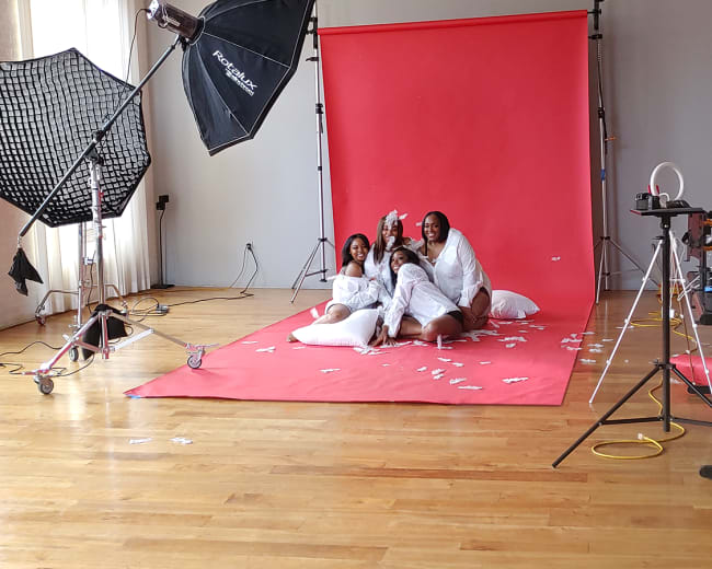 A group of four individuals in white robes poses playfully on a red backdrop in a studio setting.