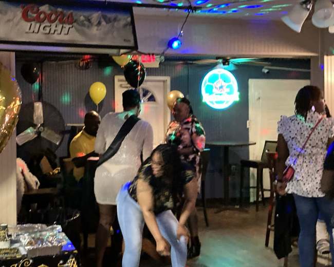A group of people socialize and dance in a brightly lit bar decorated with balloons and banners.