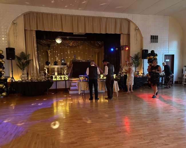 A decorated event space features a stage with a disco ball, tables adorned with decorations, and guests mingling in a ballroom setting.