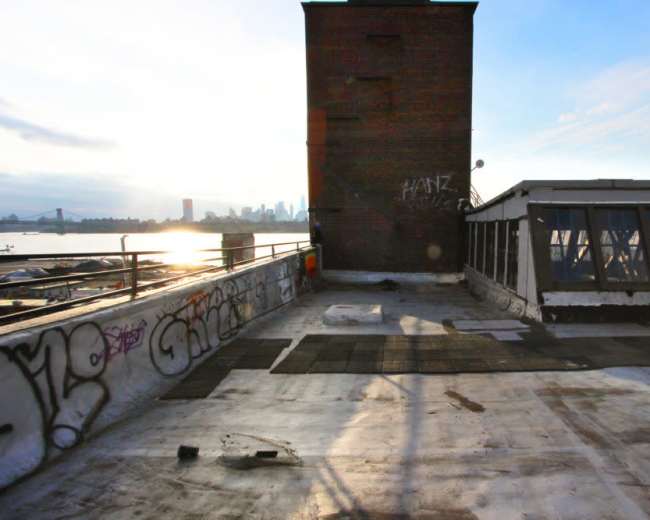 The image shows a rooftop with a brick structure and graffiti, overlooking a river and a city skyline in the background.