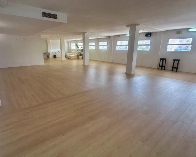 The image shows a spacious, empty room with light-colored wooden flooring and large windows, featuring minimal furniture.