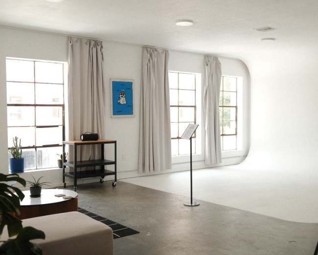 The image shows a bright, minimalistic studio space featuring large windows, a music stand, and a couch.
