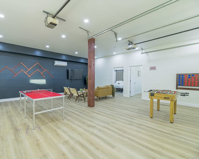 The image shows a modern recreational room featuring a ping pong table, a foosball table, seating arrangements, and wall decorations, with bright lighting and wooden flooring.