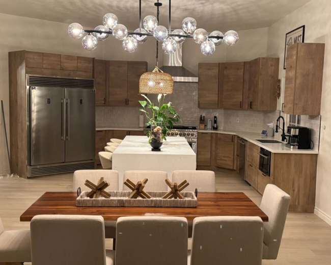 A modern kitchen features wooden cabinets, stainless steel appliances, a large island, and a dining table with decorative centerpieces.