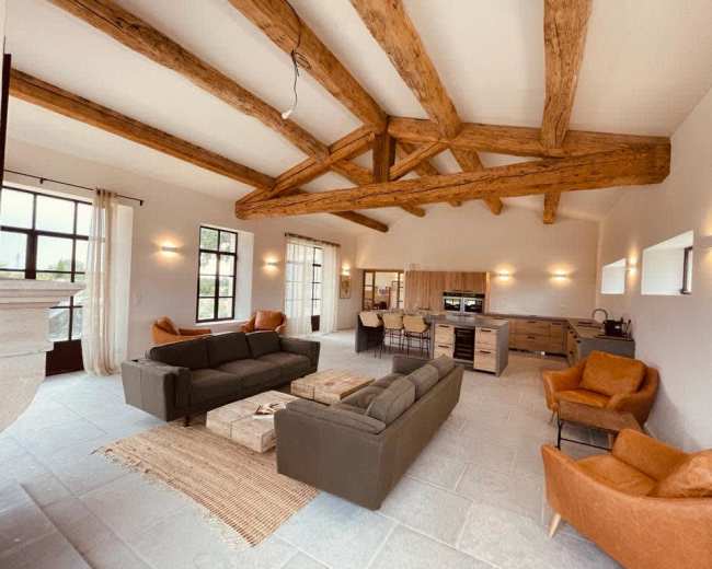 The image shows a spacious living area with exposed wooden beams, two green sofas, two brown armchairs, a coffee table, and a kitchenette in the background.