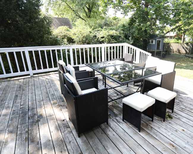 A black wicker patio set with a glass table and white cushions is arranged on a wooden deck surrounded by greenery.