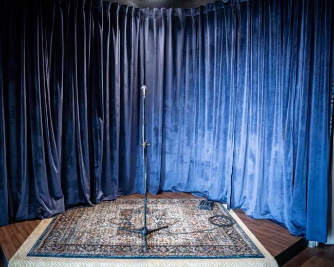 A microphone stand is positioned on a patterned rug in front of dark blue velvet curtains.