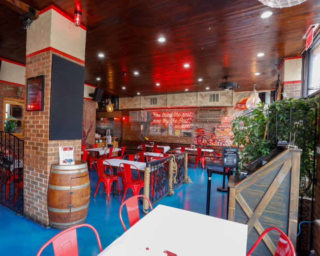The image shows a restaurant interior with brick walls, wooden ceilings, red chairs, and decorative elements including a barrel and neon signage.