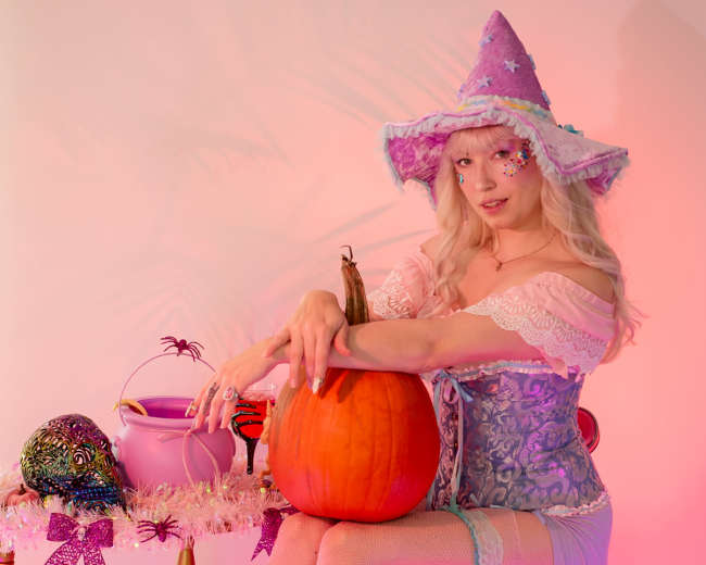 A person in a purple witch costume sits beside a pumpkin and decorative items on a small table with pink lighting.