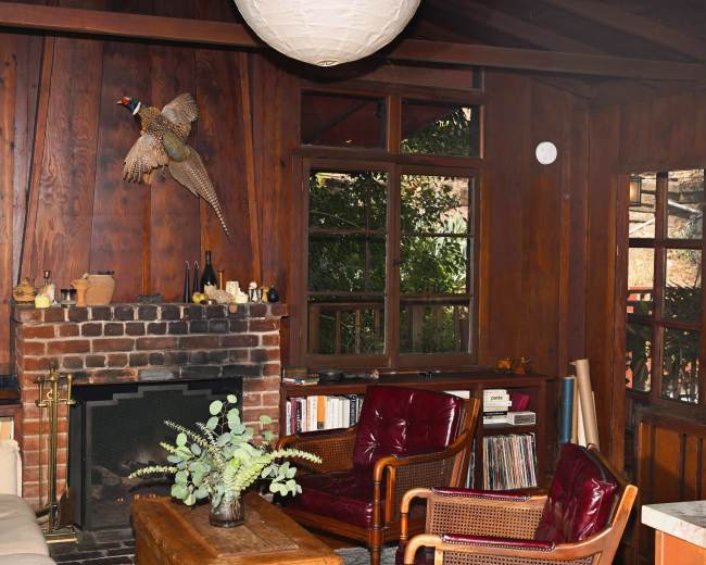 The image shows a cozy wooden interior of a room featuring a brick fireplace, a mounted bird on the wall, and comfortable seating arrangements with a coffee table.