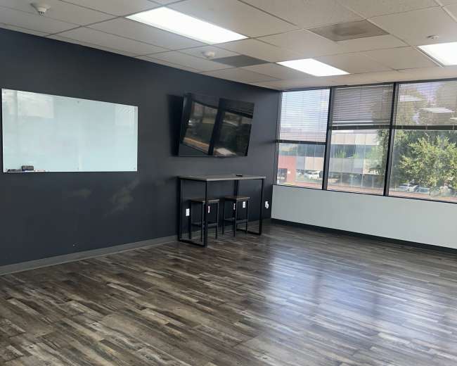 The image depicts a spacious, modern office interior with dark gray walls, wood flooring, and two flat-screen TVs mounted on the wall.