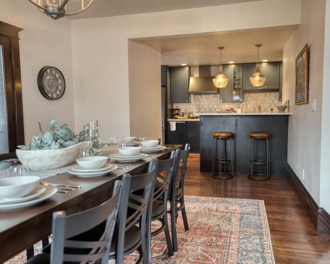 A dining area features a long table set with dishes, leading to a kitchen with dark cabinets and bar stools.