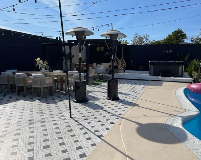 The image shows a modern outdoor space with a seating area, a pool, and two patio heaters beside a dining table.