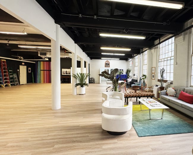 The image shows a spacious, well-lit interior with a lounge area featuring a sofa and chairs, large windows, and wooden flooring.