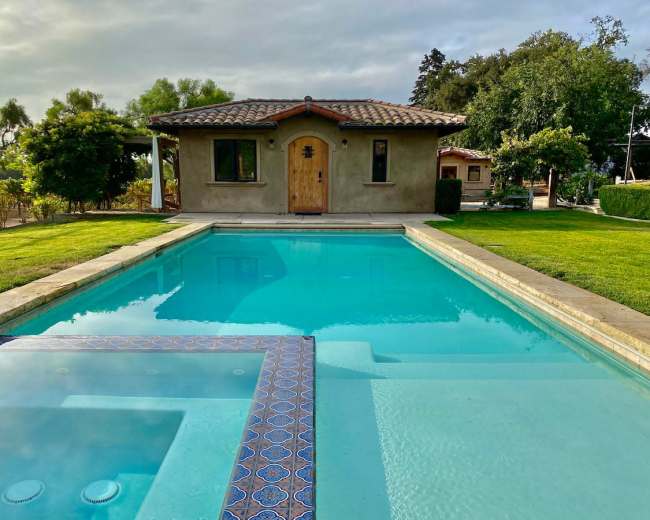 A clear swimming pool reflects a small house with a wooden door, surrounded by green grass and trees.