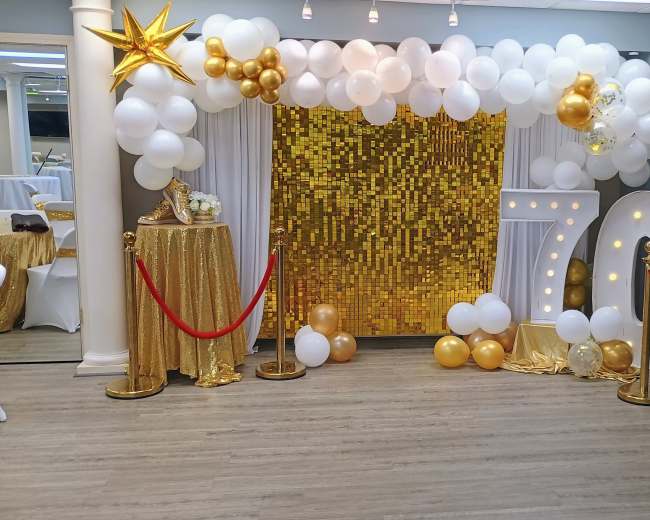 The image shows a festive event backdrop featuring a golden sequined wall, large illuminated numbers "70," and decorative balloons in white and gold, accompanied by a red carpet and stanchions.