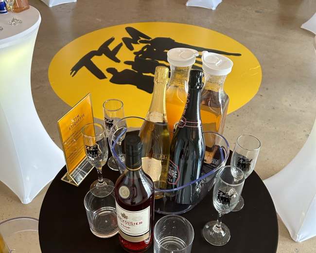 A table with a black tablecloth holds a bucket of assorted drinks, including champagne and a bottle of rum, surrounded by glassware and set in an event space with standing tables.