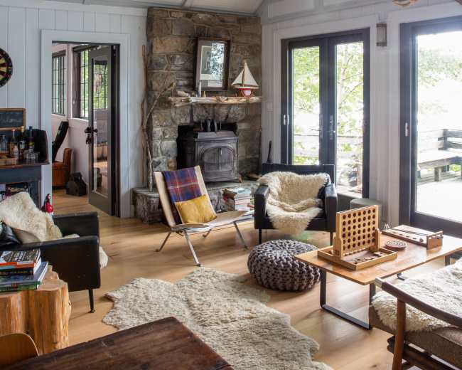 The image shows a cozy living room with a stone fireplace, large windows, and comfortable seating arrangements, including a rocking chair and a low table with a game.