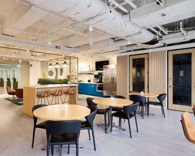The image shows a modern office interior featuring a kitchen area, dining tables, and seating spaces amidst a sleek design with open spaces and greenery.