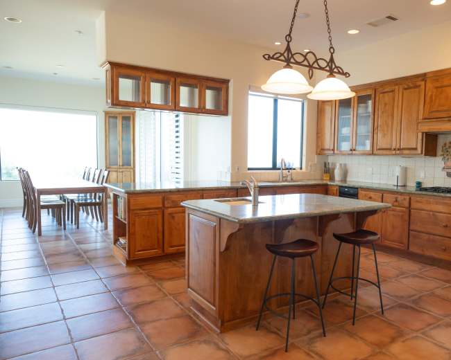 The image shows a spacious kitchen with wooden cabinetry, a large island with two stools, and a window overlooking a dining area.