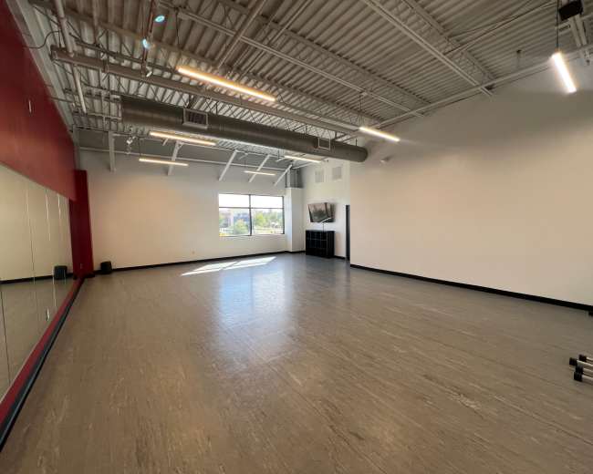 The image shows a spacious, empty fitness studio with large windows, a mirrored wall, and minimal equipment.
