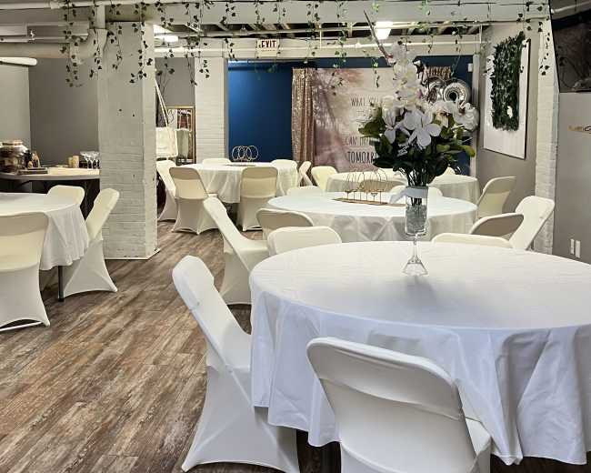 A spacious event room features white tables covered with tablecloths and surrounded by chairs, with decorative plants hanging from the ceiling.
