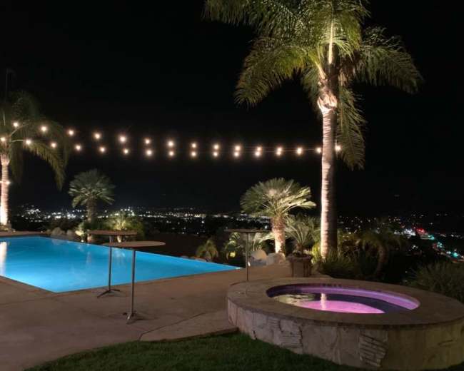 The image shows a pool area with illuminated string lights and palm trees overlooking a nighttime city view.