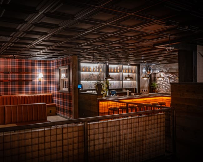 The image shows a restaurant interior with a plaid wall design, wooden accents, and a well-lit bar area.