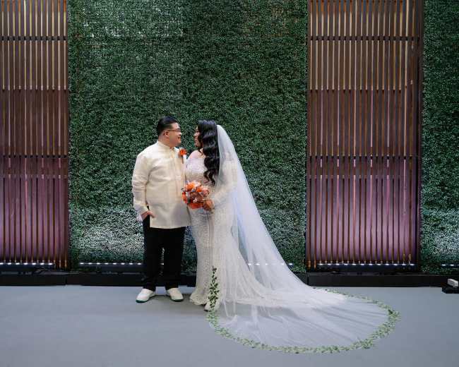 A bride in a white lace gown with a long veil stands beside a groom in a traditional barong, both smiling and holding a bouquet of orange flowers in front of a textured green backdrop.