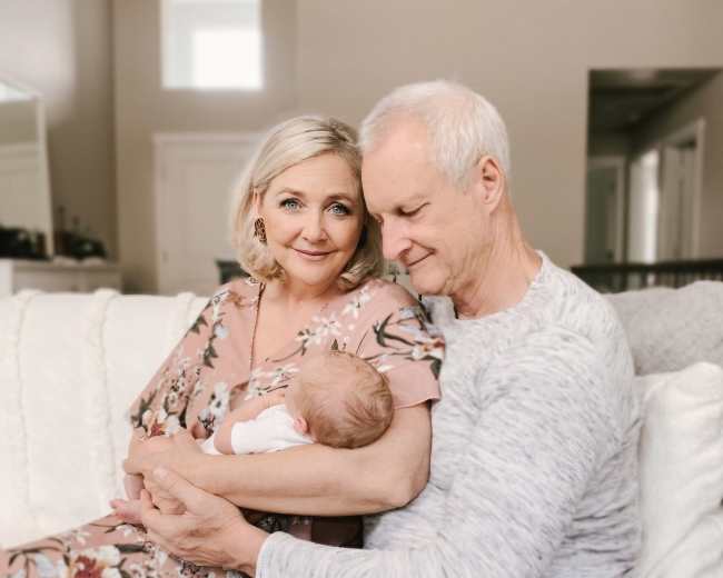 A couple sits together on a couch, holding a newborn baby in their arms, with soft natural light illuminating the room.