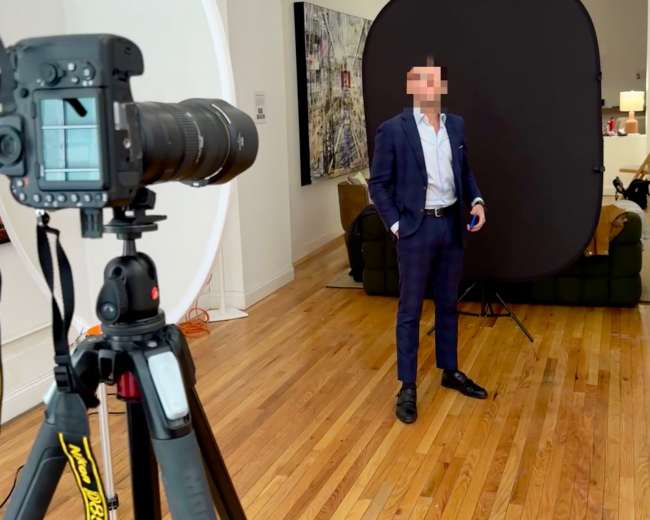 A man in a blue suit stands in front of a black backdrop while a camera on a tripod points towards him.