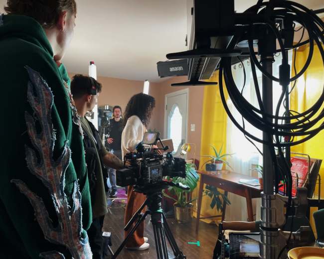 A group of people is filming a scene in a well-lit room with cameras and lighting equipment set up.