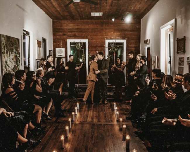 A couple stands facing each other and embracing at the front of a candlelit room filled with seated guests who are applauding.