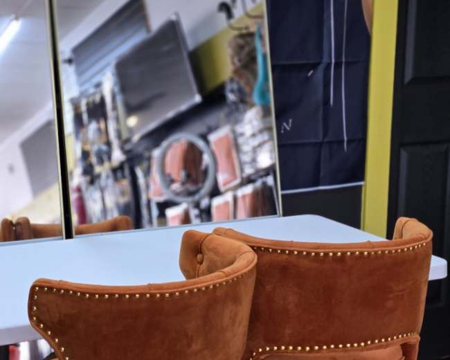 The image shows two orange velvet chairs with studded detailing positioned at a white table in a store setting that features mirrors and decorative items in the background.