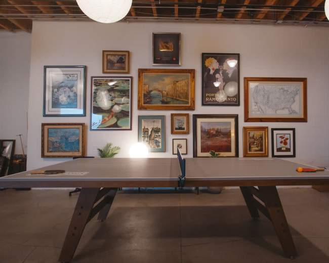 A ping pong table is positioned in front of a wall adorned with various framed artworks and photographs in an indoor space.