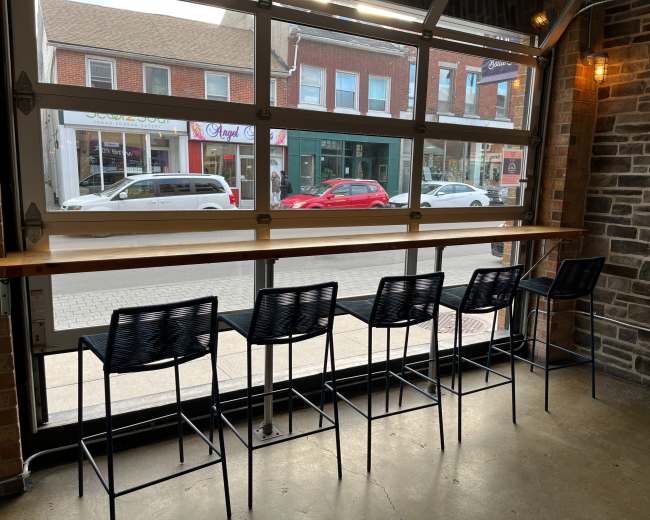 The image shows a row of five black bar stools at a wooden counter next to a large window, revealing a street view with parked cars and storefronts.