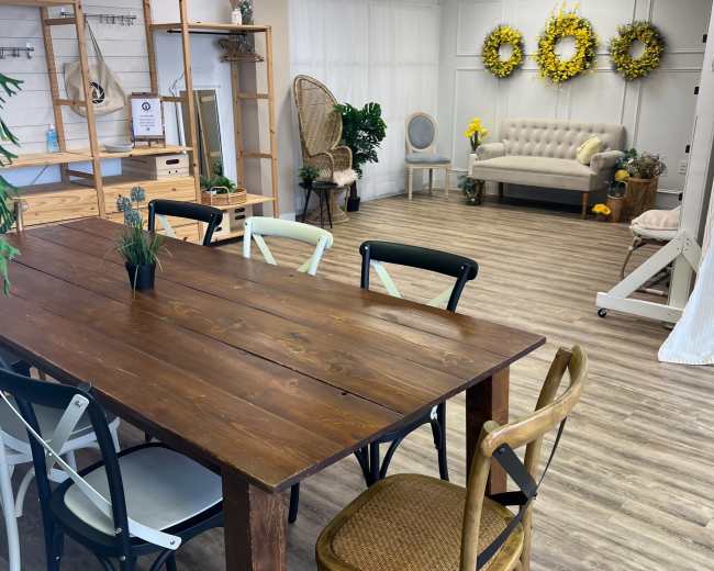 The image shows an indoor space with wooden flooring, featuring a large wooden dining table surrounded by various chairs, a cozy sofa, and decorative elements like sunflowers on the walls.