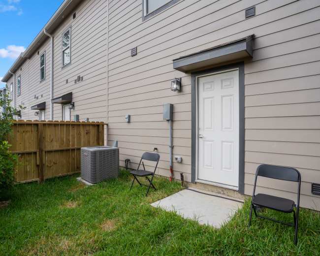 The image shows a small backyard area with a door, a concrete slab, two folding chairs, a utility box, and an air conditioning unit beside a wooden fence.