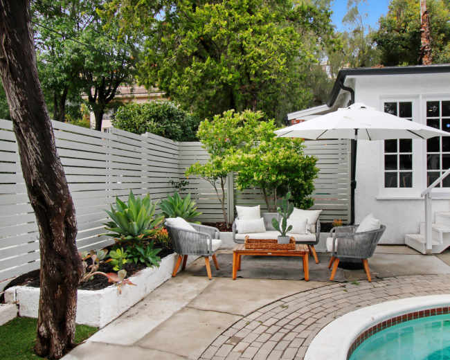 A cozy outdoor seating area features two chairs and a table under a white umbrella, adjacent to a pool and surrounded by plants and a white fence.