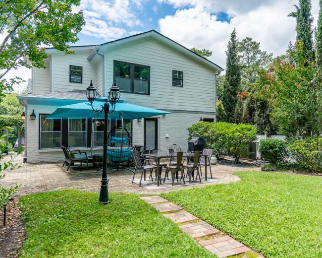 The image shows a backyard patio with a seating area, a table with chairs, and a large umbrella, surrounded by grass and landscaped trees.