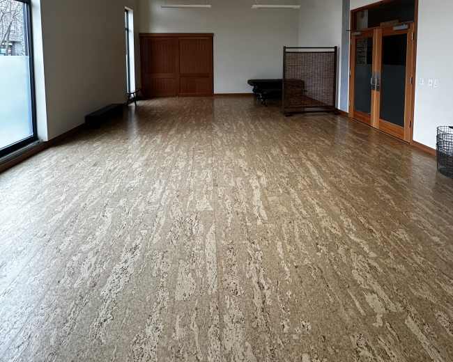 The image shows a spacious, empty room with large windows, wooden ceiling fans, and a wooden sliding door on one side.