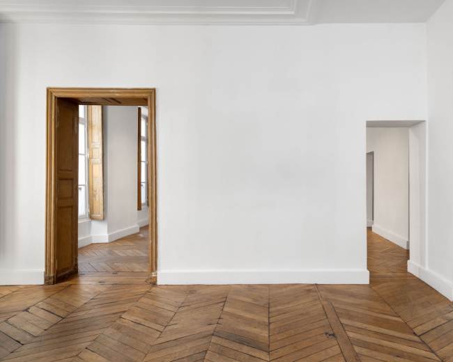The image shows a minimalist room with white walls, wooden herringbone flooring, and two doorways leading to other spaces.
