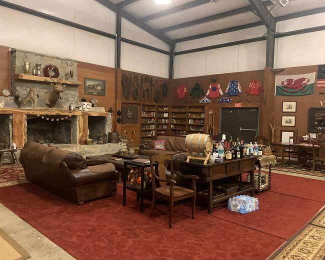 The image shows a spacious rustic room with a seating area featuring leather sofas, a large stone fireplace, and various decorations, including mounted animal heads and shelves filled with bottles and memorabilia.