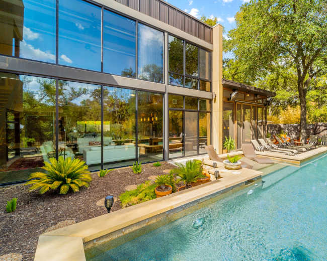 The image shows a modern house with large glass windows overlooking a swimming pool surrounded by landscaping and lounge chairs.
