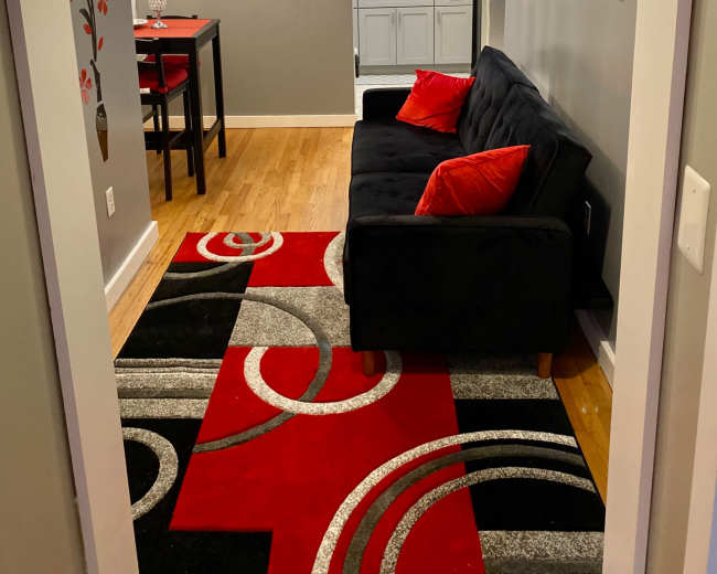 The image shows a small living area featuring a black sofa with red cushions, a patterned rug, and a view of a kitchen area with modern cabinetry and a dining table.