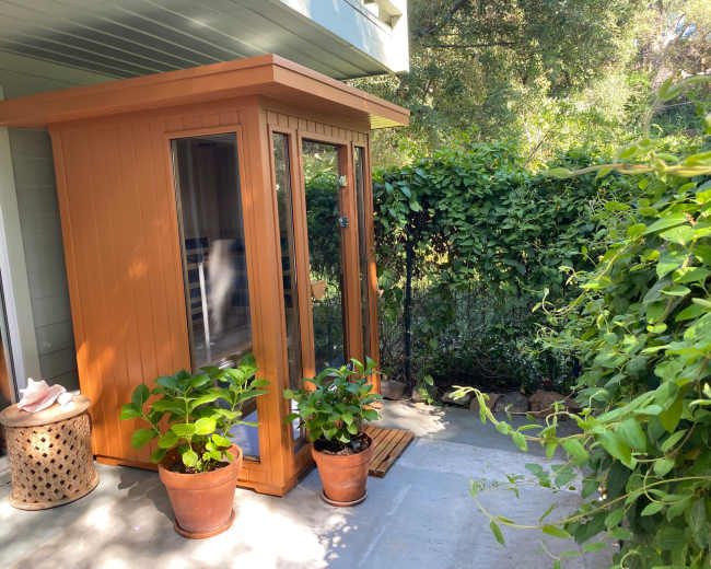 The image shows a small orange garden shed with glass doors surrounded by greenery and potted plants on a concrete patio.