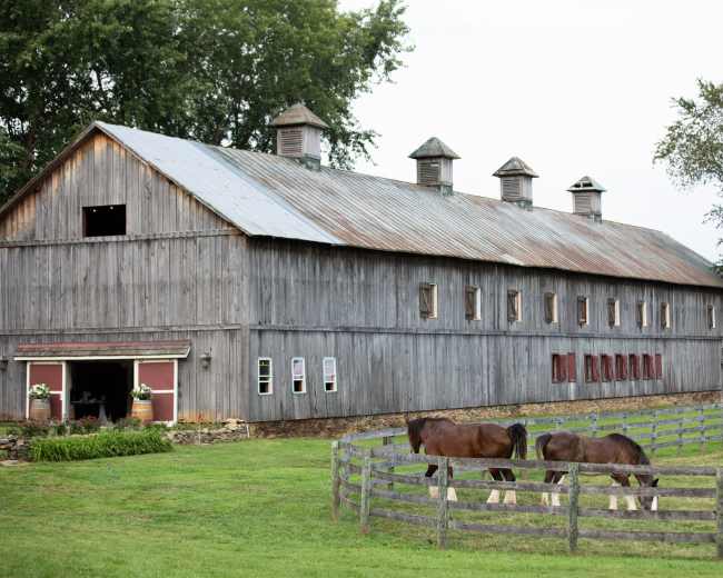 A weathered wooden barn stands beside a fenced pasture, where two horses graze in the grass.