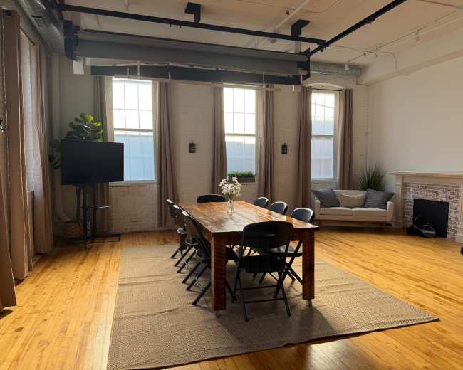 The image shows a brightly lit interior space with a long wooden table surrounded by black chairs, situated on a textured rug, and features large windows with curtains, a sofa, and decorative plants.